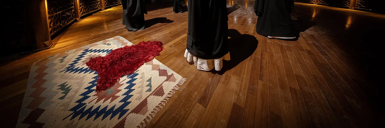 Uma pele de carneiro vermelha sobre um kilim estampado num piso de madeira, com os mantos escuros e as barras brancas das vestes de semazens em p&eacute; na borda da imagem