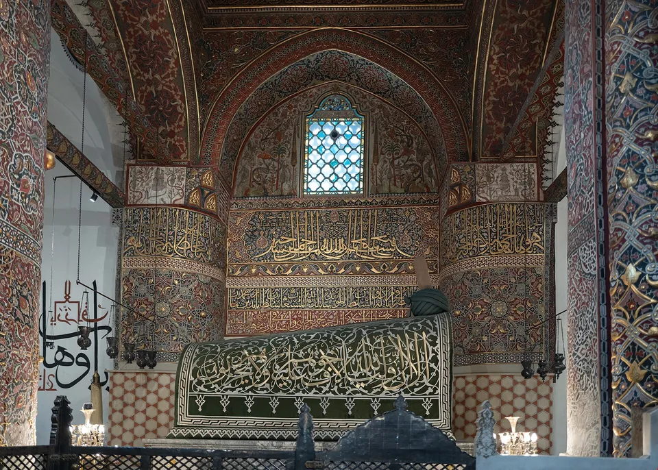Um t&uacute;mulo sob um tecido verde-escuro bordado com escrita &aacute;rabe, numa sala com paredes e arcos pintados com ornamentos e caligrafia, atr&aacute;s de uma grade de metal