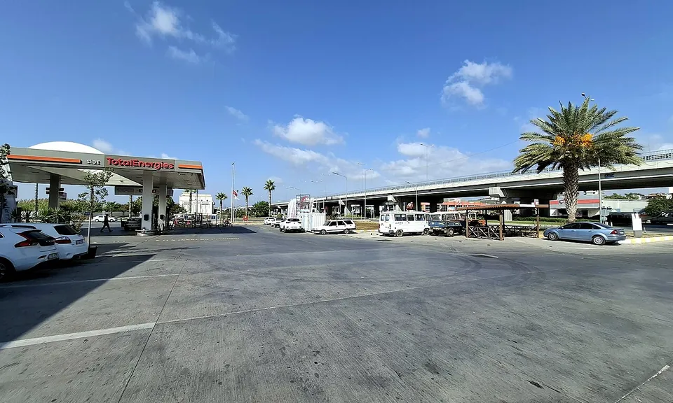 Une vaste aire de station-service au soleil, avec un palmier et un pont routier derrière, quelques voitures garées