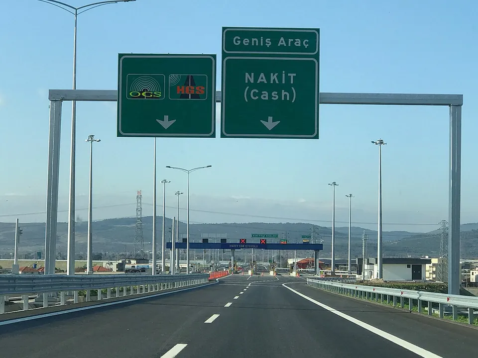 Un portique au-dessus d’une autoroute à péage avec deux panneaux verts de voies : l’un avec les symboles OGS et HGS, l’autre portant Geniş Araç au-dessus de Nakit (espèces)