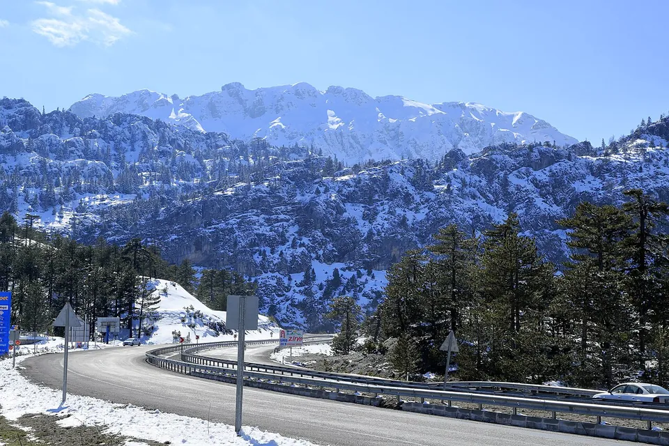 Une route sinueuse bordée d’une glissière de sécurité traversant une forêt de pins sous des montagnes enneigées, avec une voiture et des panneaux dans le virage
