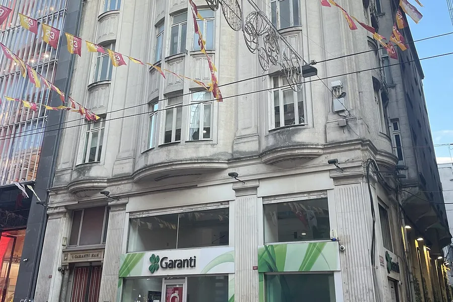 İstanbul'da kalabalık bir yaya caddesinde, zemin kattaki girişinin &uuml;zerinde yeşil Garanti tabelası bulunan taş bir k&ouml;şe bina; caddenin &uuml;zerine bayraklar asılmış