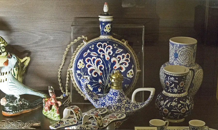 Стъклена витрина със синьо-бяла рисувана керамика: кръгла бутилка с тапа, кани, чаши и малки фигурки върху дървен рафт