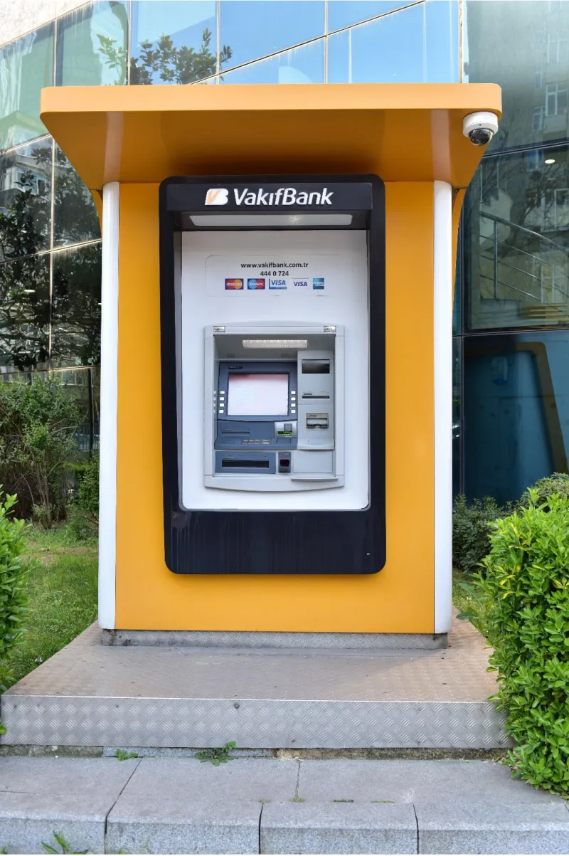 A yellow cash machine in a roofed kiosk in front of a glass building in Istanbul, with a row of card-network logos above its screen