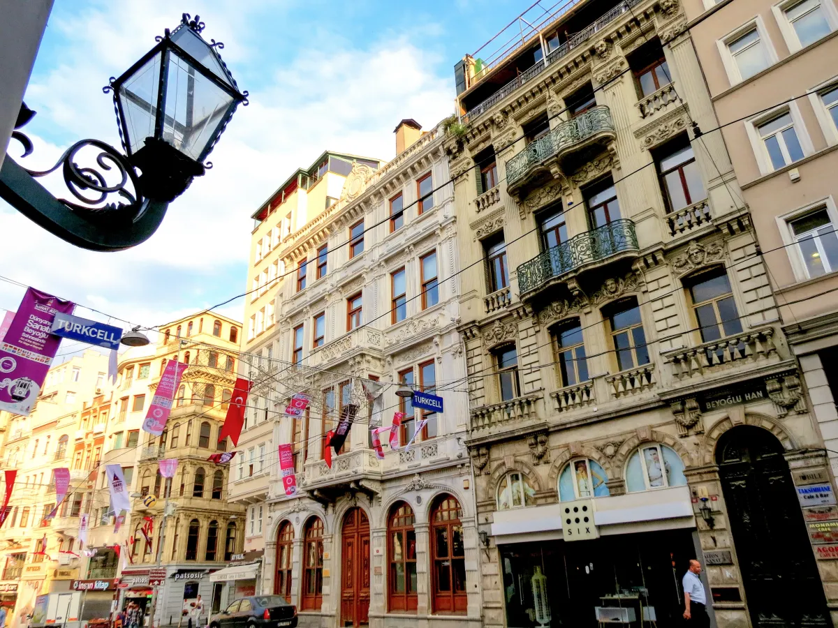 A pedestrian street in Istanbul lined with stone apartment buildings, shop signs and hanging banners, seen from below with a street lamp in the corner
