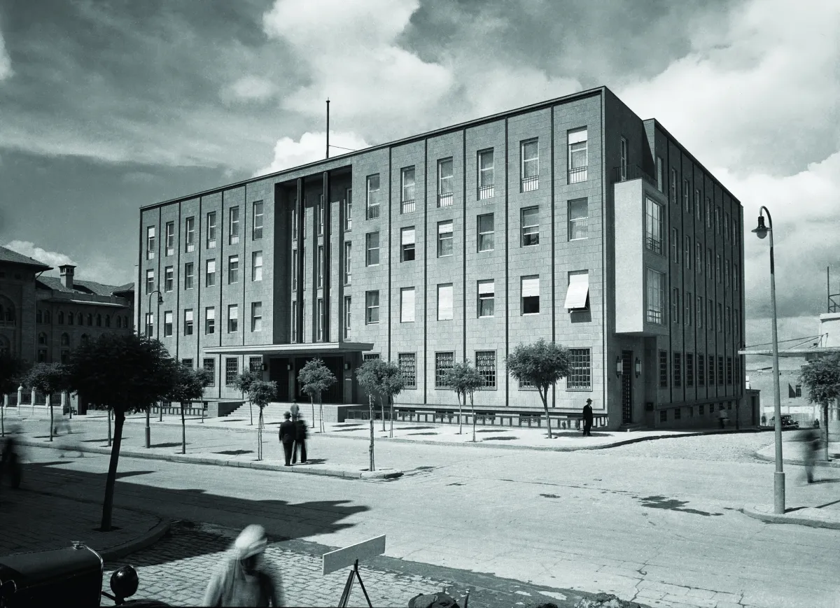 A black-and-white photo from the 1940s of a four-storey stone building with a tall column of windows over its entrance, on a tree-lined Ankara street with a few people and a car