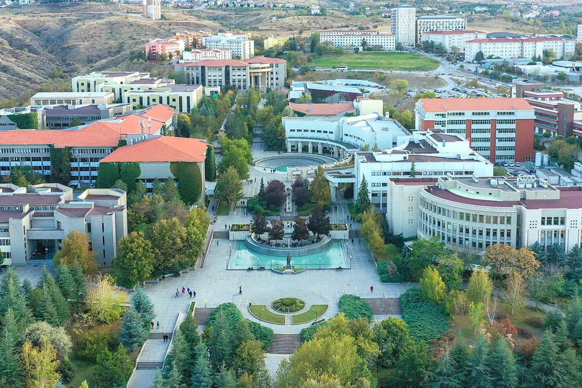 俯瞰一座大学校园:红顶建筑和白色楼宇环绕着带喷泉的水池,四周有松树和秋色中的树木,后方是干燥的山丘。