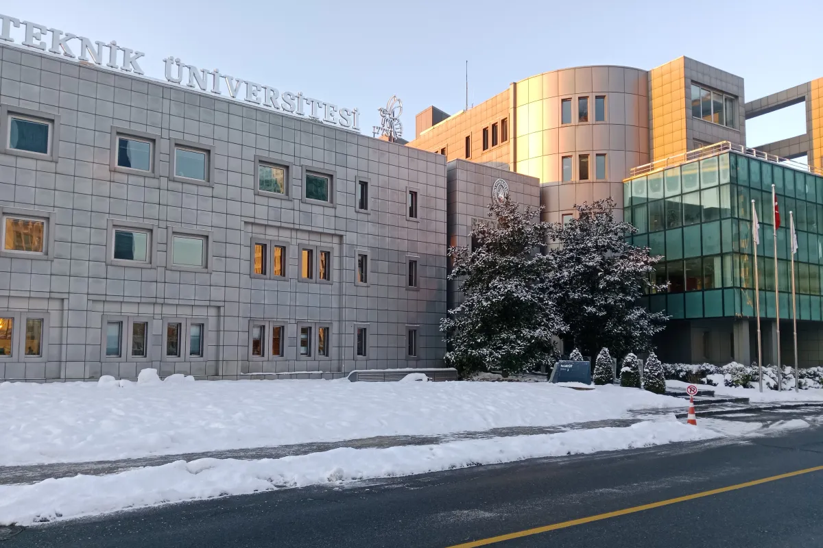 一座灰色石材外墙的大学建筑,屋顶立着大字TEKNİK ÜNİVERSİTESİ,并饰有蜜蜂徽标,旁边是由绿色玻璃和铜色面板组成的楼体,前方有三根旗杆。地面和树木上覆盖着积雪,画面中无人。