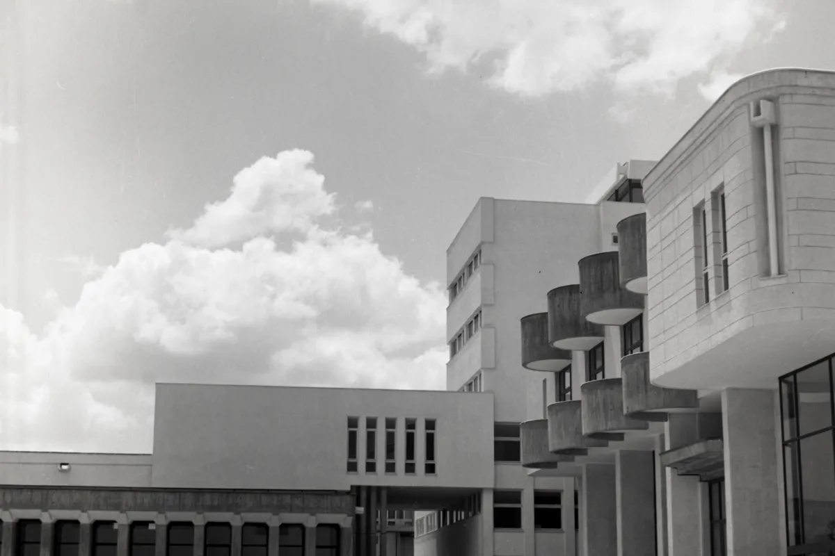 现代大学建筑的黑白照片:转角塔楼上层层叠起圆形混凝土阳台,旁边是一座石材外墙楼体和开有窄窗的平顶侧翼。上方是一大片云,画面中无人。
