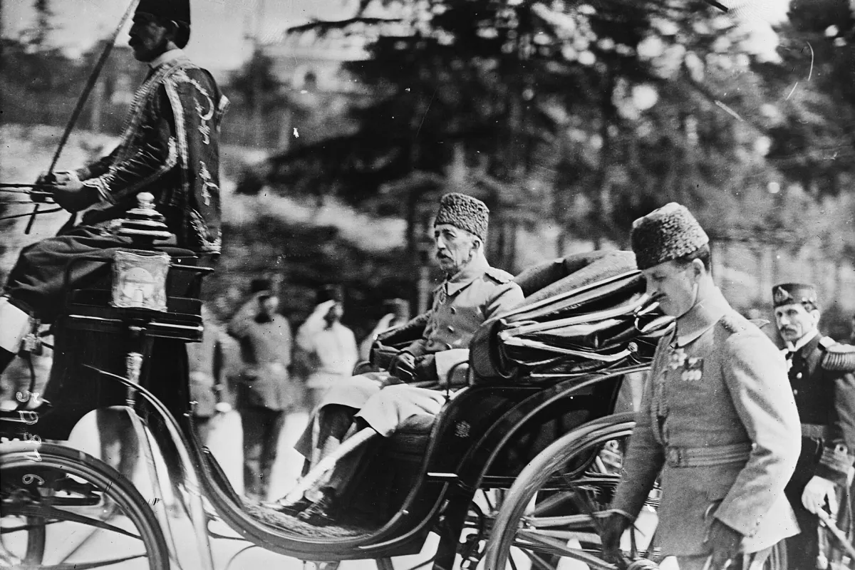 Mehmed VI. sitzt in Uniform und mit Pelzmütze in einer offenen Kutsche, die ein Kutscher in einem bestickten Mantel lenkt; ein Offizier geht daneben