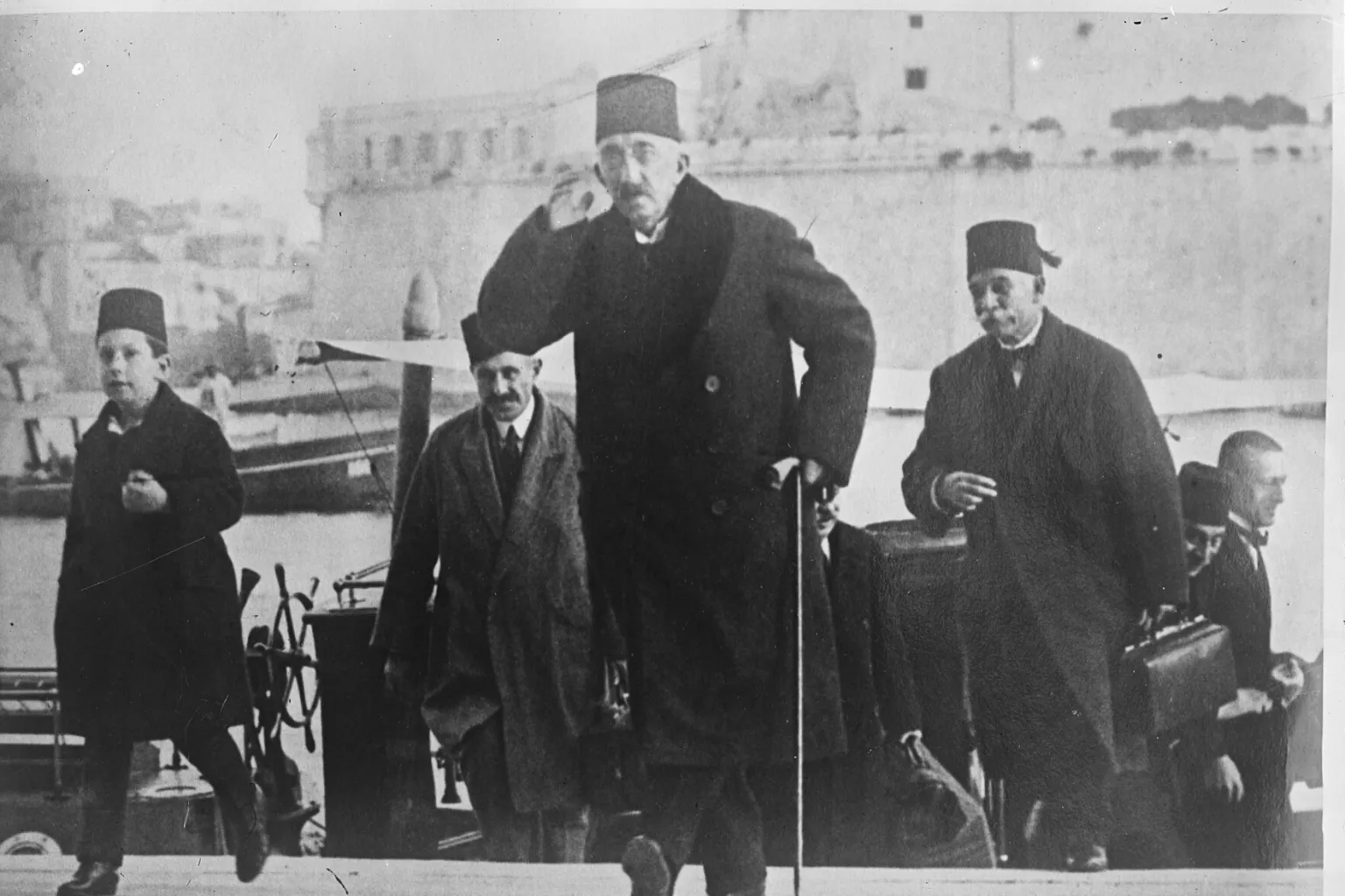 Mehmed VI. mit Fes und langem Mantel hebt die Hand an seinen Fes, als er in Malta einen Kai betritt; links geht ein Junge mit Fes, dahinter folgen zwei ältere Männer