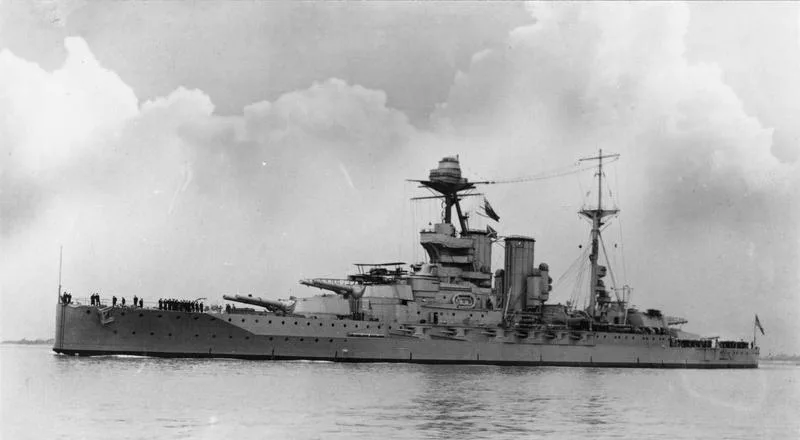 Ein graues Dreadnought-Schlachtschiff mit zwei Schornsteinen, Dreibeinmasten und großen Geschütztürmen fährt vorbei; die Besatzung steht aufgereiht an Deck