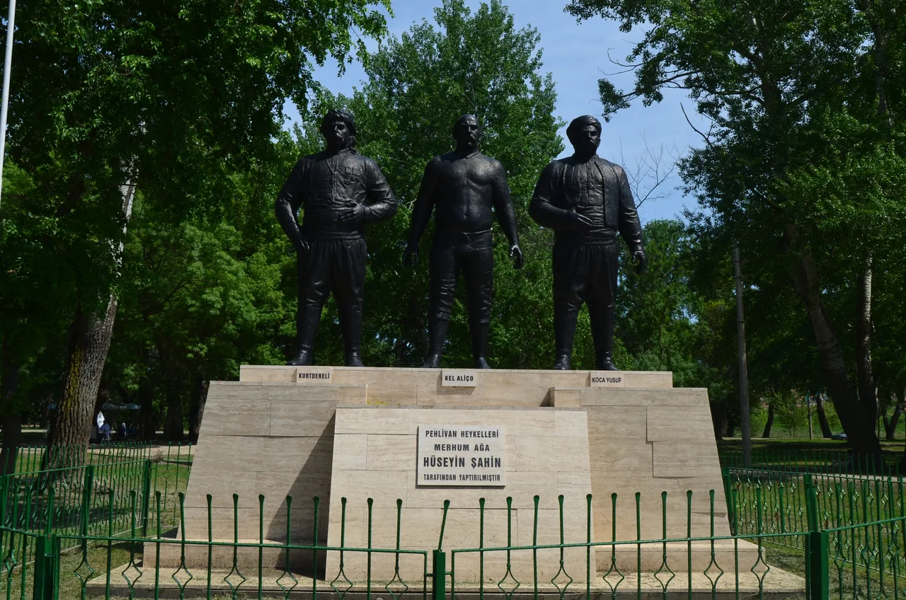 Trois statues en bronze de lutteurs debout sur un socle en pierre parmi les arbres&nbsp;; le socle porte les noms de Kurtdereli, Kel Ali&ccedil;o et Koca Yusuf