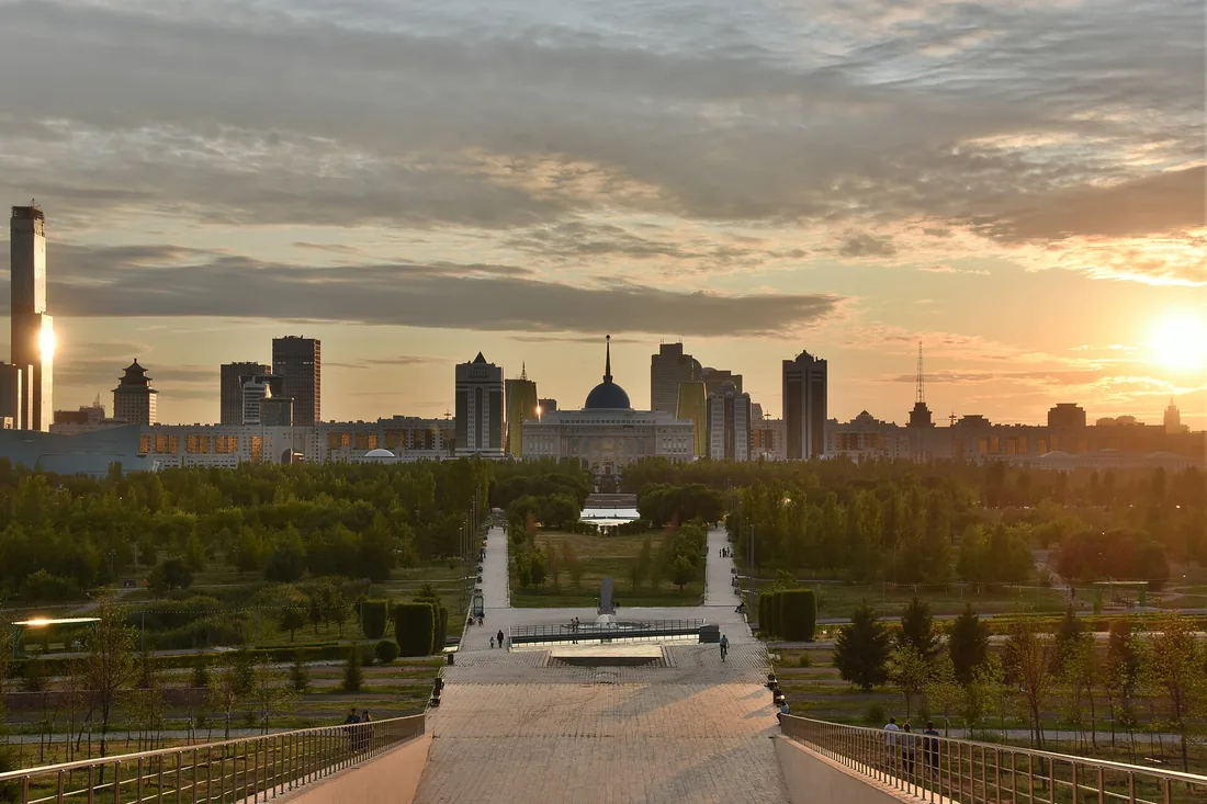 Astana o zachodzie słońca: brukowana promenada przez park prowadzi ku panoramie wieżowc&oacute;w i budynkowi z kopułą