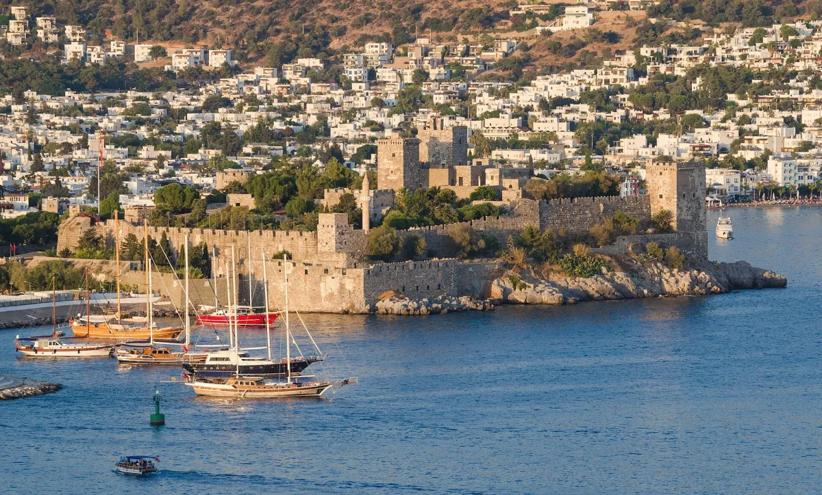 El castillo de Bodrum en una punta rocosa sobre una bahía azul, con goletas amarradas delante y casas blancas que ascienden por la colina detrás