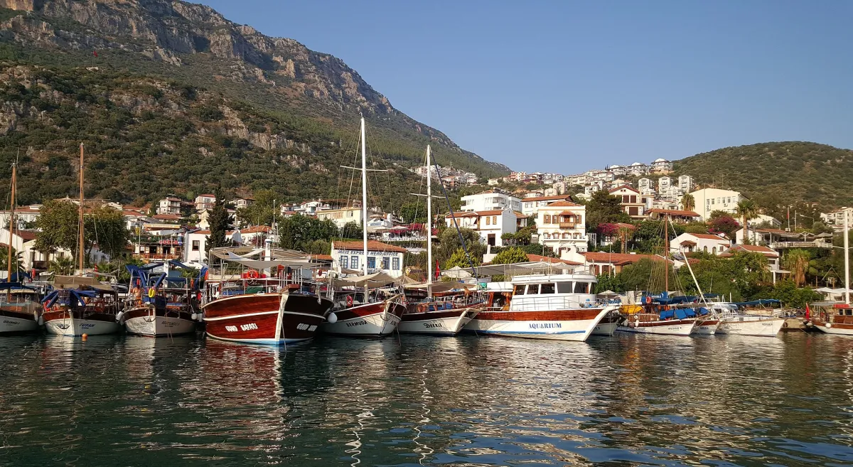 Un puerto de Kaş con goletas de madera amarradas frente a casas blancas de tejados rojos y una ladera verde bajo un cielo despejado