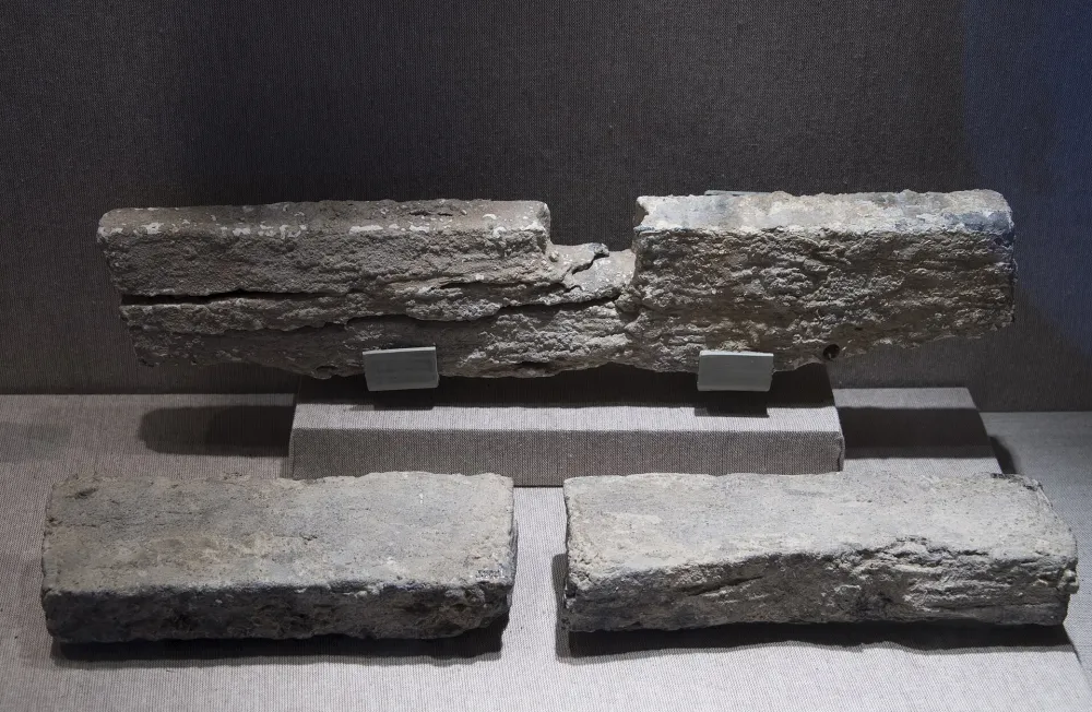 Tres piezas de plomo grises sobre una base de exposición clara: una pieza larga en un soporte y dos más cortas delante