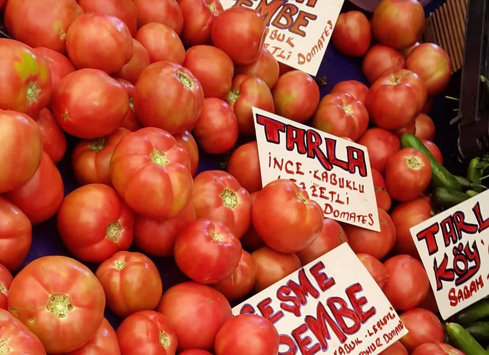 Pomodori rossi ammucchiati su un banco con cartellini scritti a mano: Tarla İnce Kabuklu Domates, Çeşme Pembe e Tarla Köy