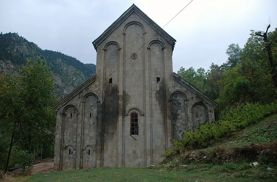 Una iglesia de piedra gris con fachada alta a dos aguas e hileras de hornacinas estrechas en arco, entre &aacute;rboles en una ladera verde