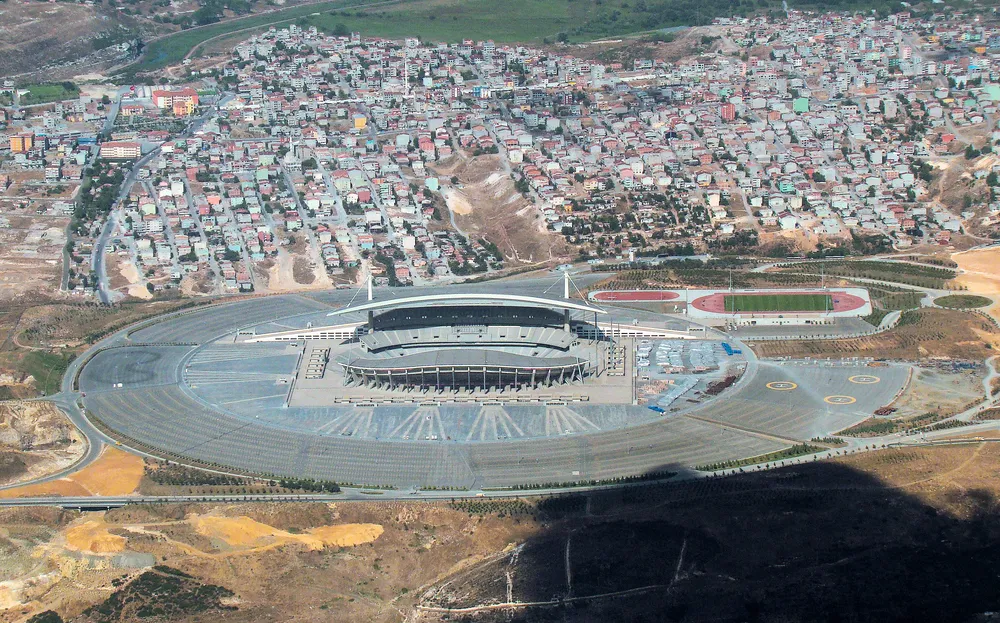 Une vue aérienne d’un grand stade ovale entouré d’un anneau de dallage gris, avec une piste d’athlétisme et un terrain d’entraînement à côté, un quartier dense de maisons à l’arrière et des collines dénudées en contrebas