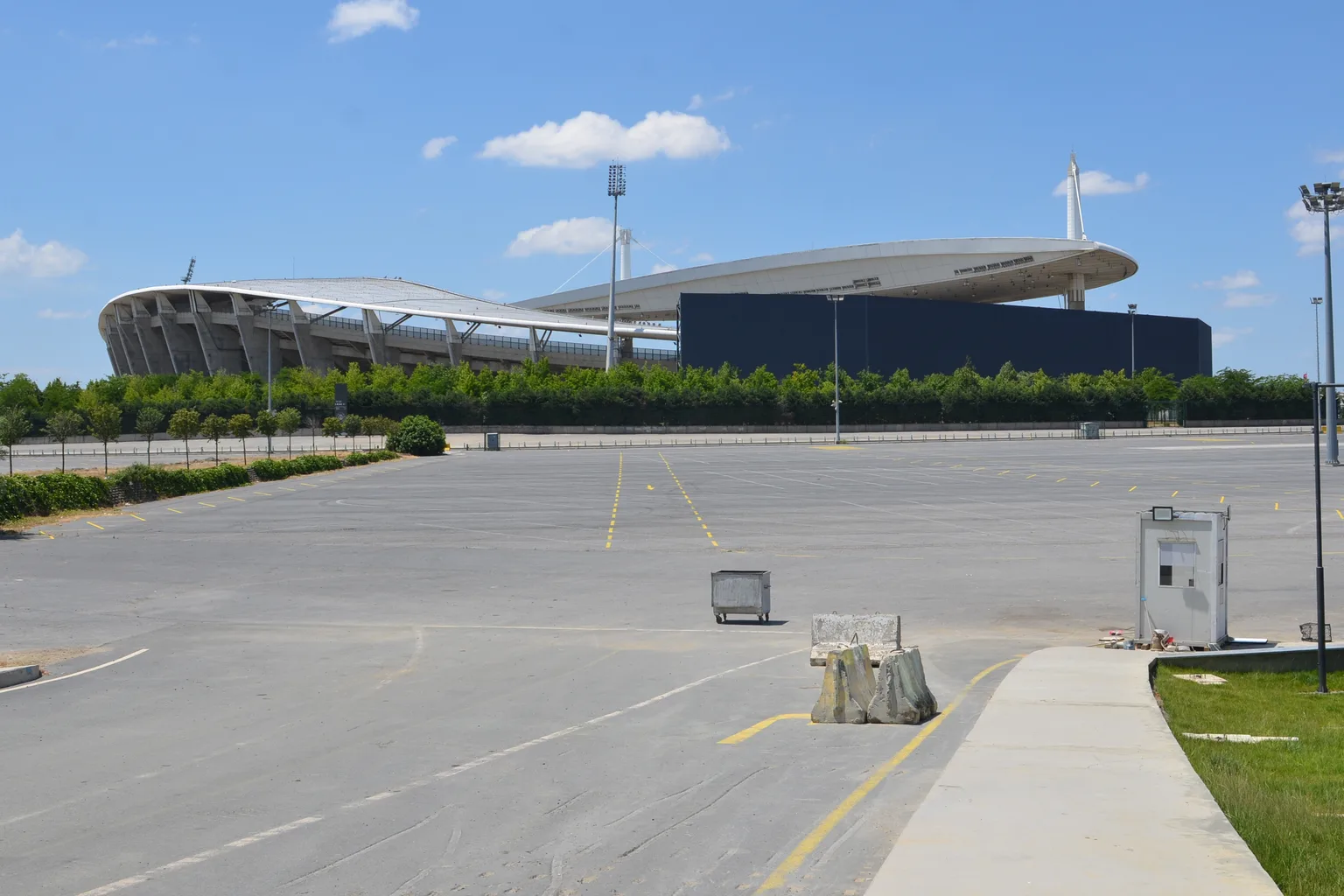 Un long toit de stade blanc avec un mât et un mur sombre à l’arrière, vu depuis un parking asphalté vide sous un ciel bleu