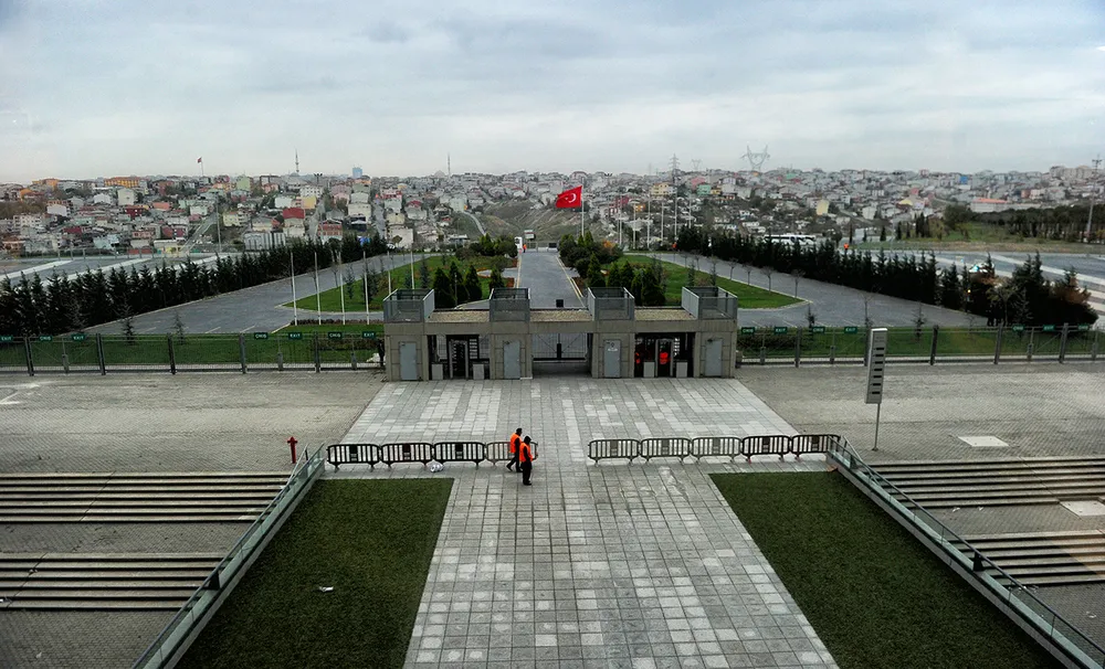 Une large allée pavée entre deux pelouses mène à une porte en pierre, avec un drapeau rouge à l’arrière, une rangée de barrières métalliques et deux stewards en gilet orange ; des maisons se dressent sur la colline au loin