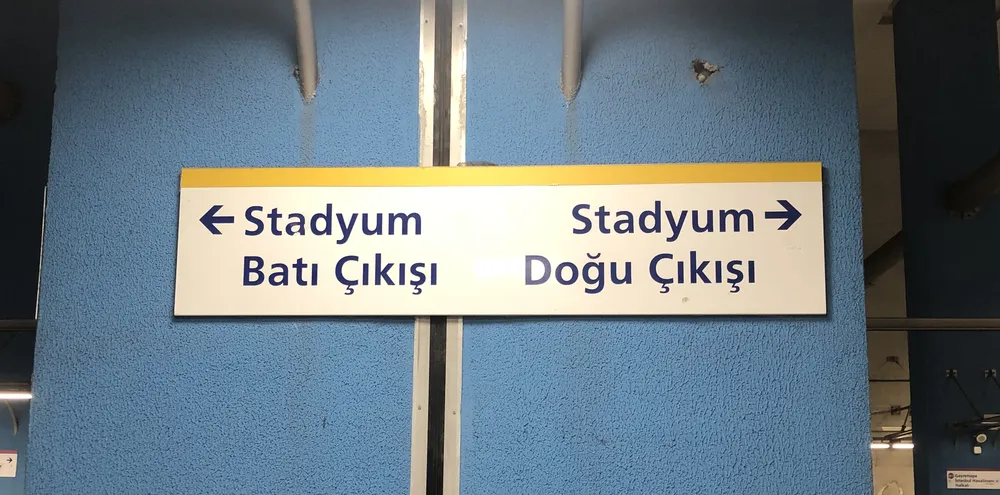 Un pilier bleu dans une station de métro, avec un panneau blanc indiquant Stadyum Batı Çıkışı et une flèche vers la gauche, et Stadyum Doğu Çıkışı et une flèche vers la droite