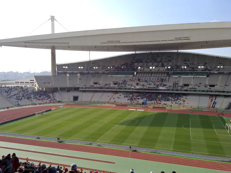 Un vaste terrain de football entouré d’une piste d’athlétisme, un long toit de tribune blanc soutenu par un mât et des rangées de sièges avec quelques spectateurs