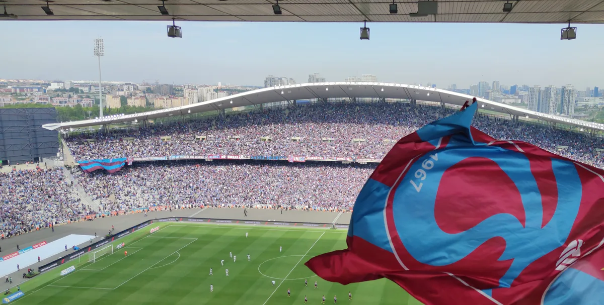 Un stade de football plein vu des tribunes supérieures : un long toit incurvé au-dessus des gradins remplis en face, un terrain vert avec des joueurs et un grand drapeau rouge et bleu au premier plan