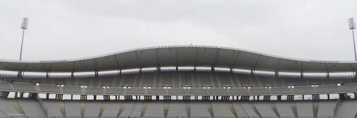 Un long toit de stade blanc ondulé au-dessus de rangées de sièges gris, vu depuis l’autre côté d’un terrain sous un ciel pâle, avec un mât de projecteurs à chaque extrémité