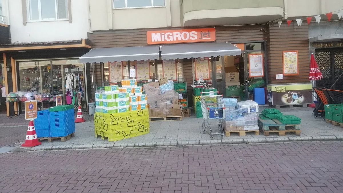 Uma pequena loja Migros Jet em uma rua lateral, com toldo listrado, caixas e produtos empilhados na calçada em frente