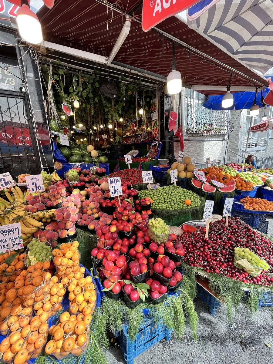 Uma banca de hortifrúti repleta de laranjas, maçãs, cerejas, uvas e melões sob luminárias penduradas, com cartões de preço escritos à mão
