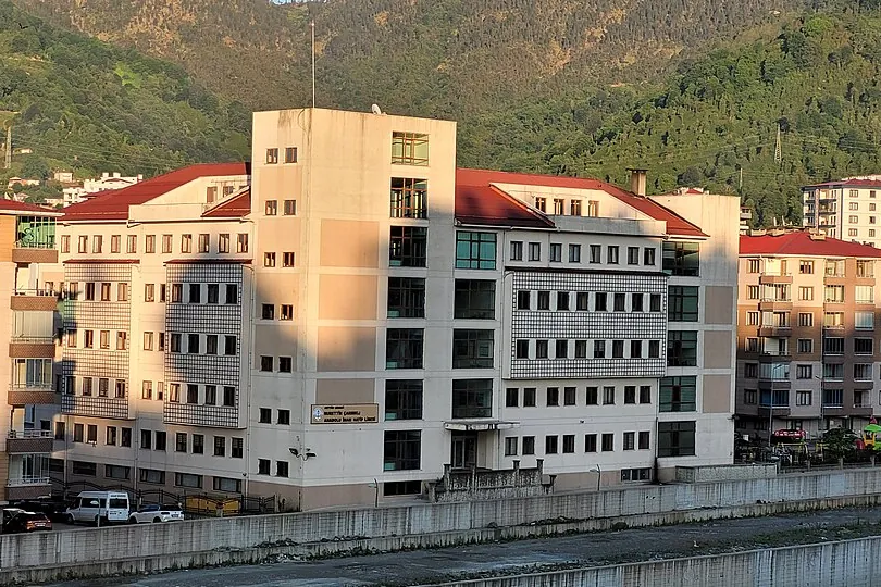 Um bloco escolar moderno de v&aacute;rios andares junto a um rio com muro de conten&ccedil;&atilde;o e uma colina verde ao fundo
