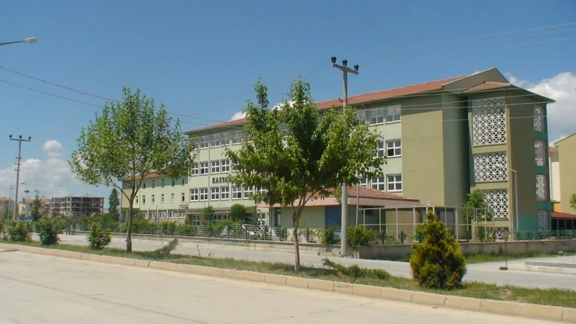 Um bloco escolar verde e branco com telhado de telhas numa rua tranquila, &aacute;rvores jovens &agrave; frente e um poste de energia junto &agrave; via