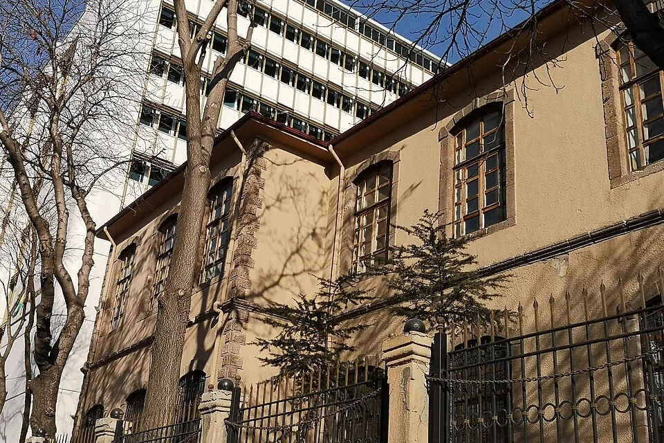 Um antigo muro escolar ocre com janelas em arco e uma cerca de ferro ao lado de um bloco moderno mais alto, com &aacute;rvores sem folhas &agrave; frente