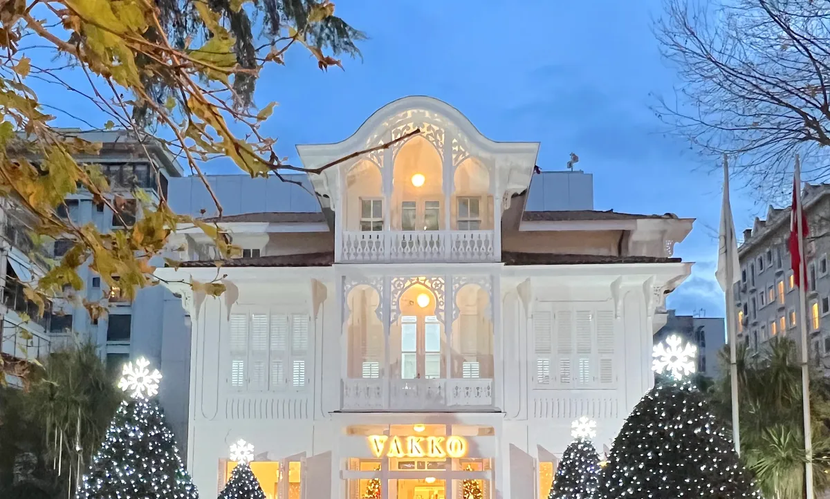 Una mansi&oacute;n blanca de madera en Estambul iluminada al anochecer, con VAKKO en letras doradas sobre la puerta y &aacute;rboles iluminados a ambos lados