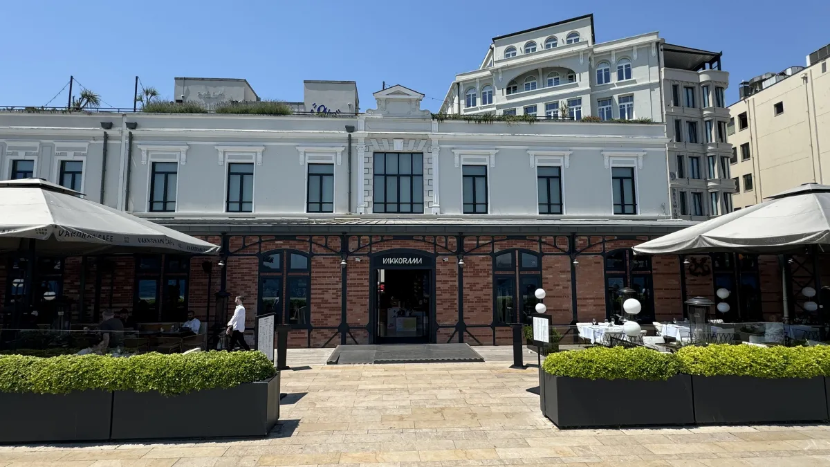 Edificio revocado con una base de ladrillo frente al mar en Galataport, con VAKKORAMA sobre la puerta y mesas de caf&eacute; bajo sombrillas grises