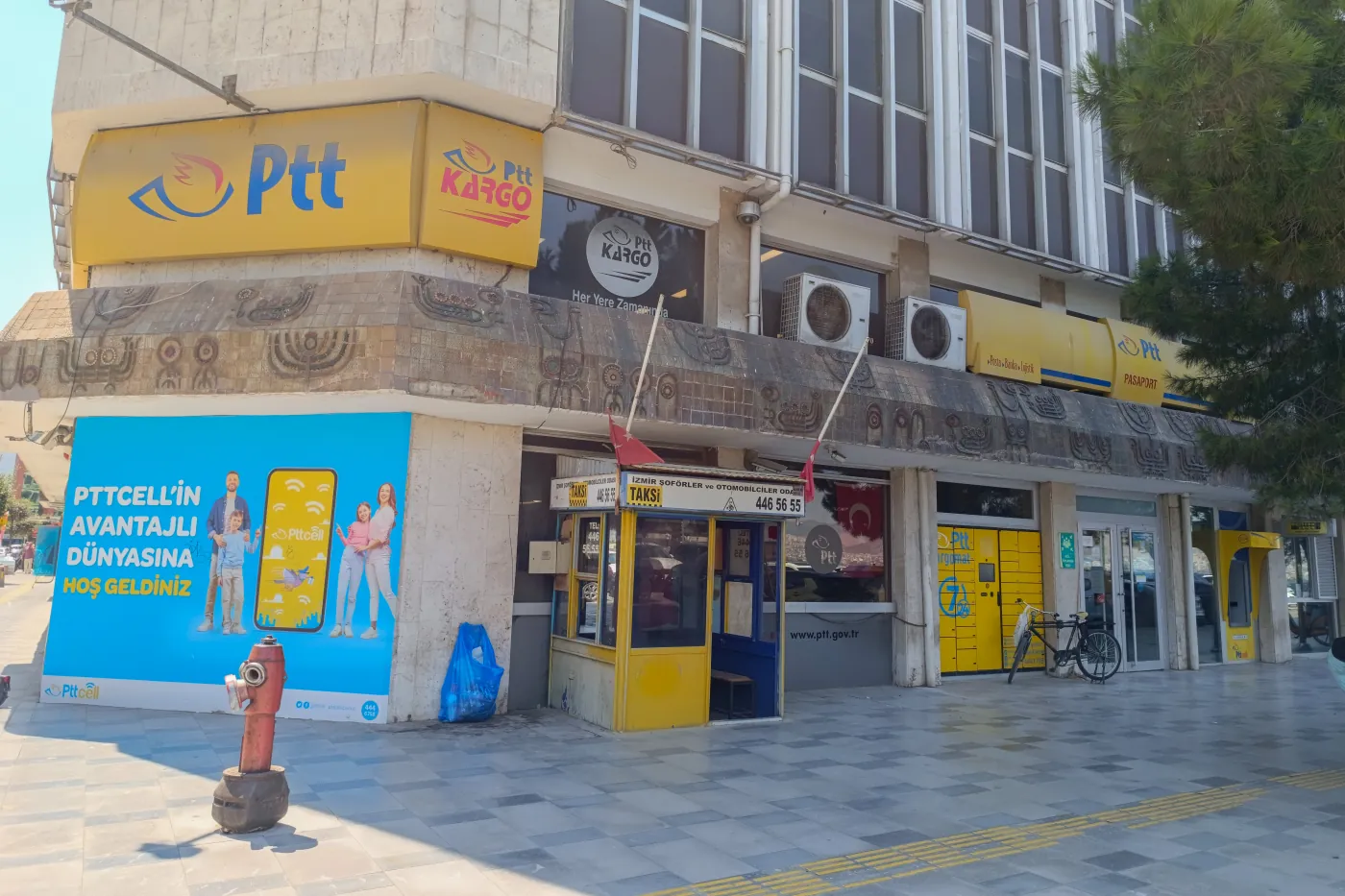Un bureau de poste à l’angle d’une rue, avec de grandes enseignes jaunes Ptt et Ptt Kargo et une rangée de consignes à colis jaunes à côté de l’entrée.