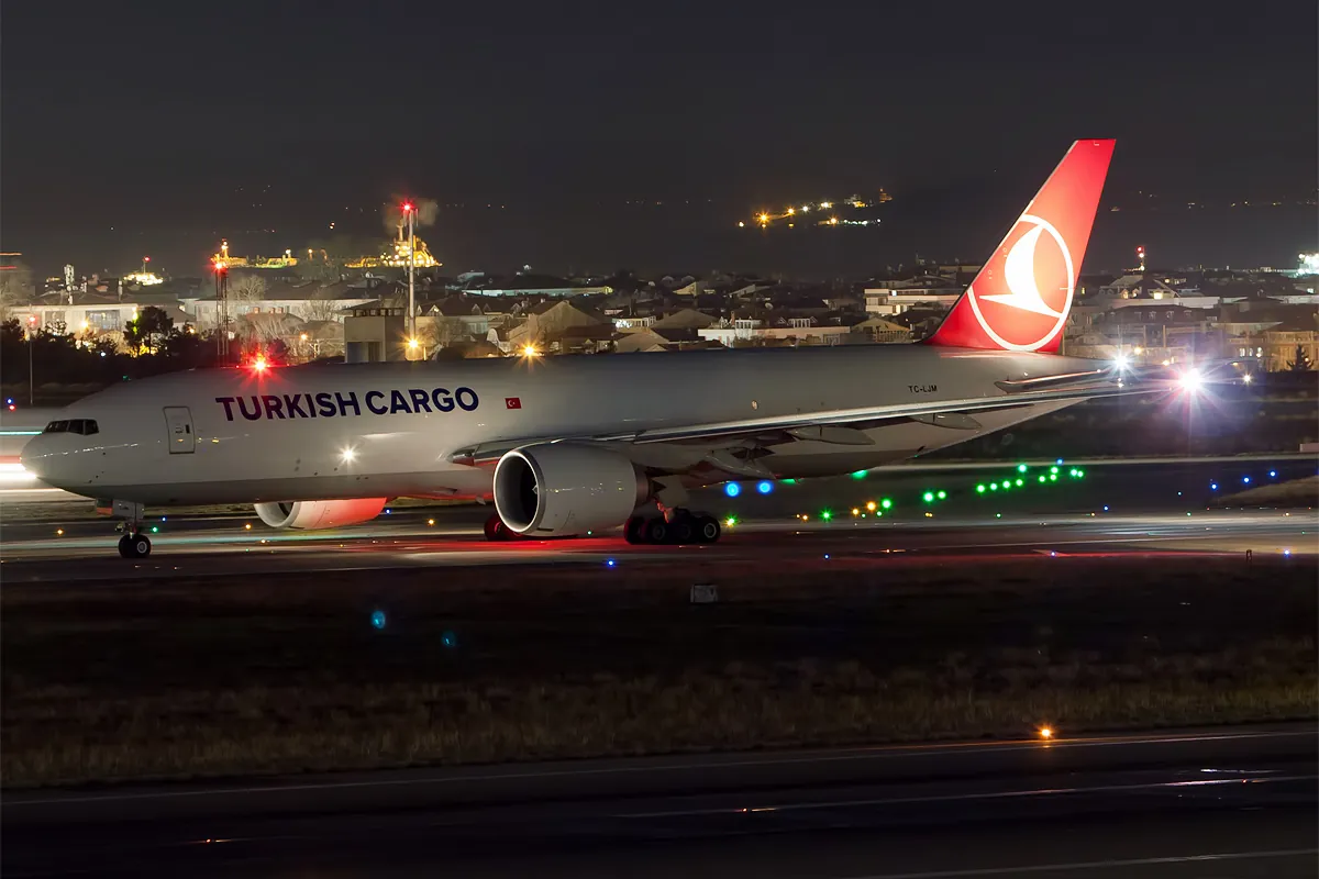 Un avion-cargo Boeing 777 de Turkish Cargo avec une dérive rouge dans un aéroport de nuit, entouré de feux de piste.
