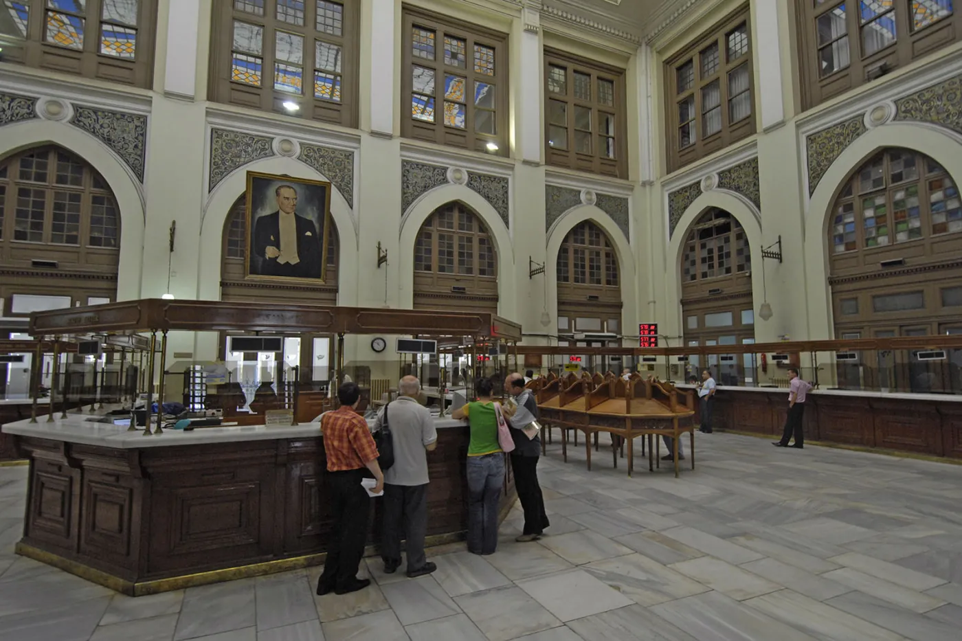 Des personnes aux guichets en bois dans le haut hall central d’un grand bureau de poste, avec des fenêtres cintrées.