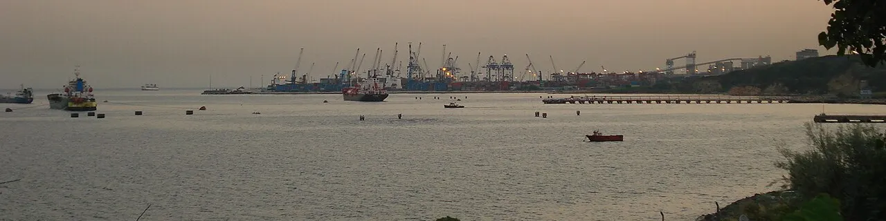 Foto: i terminal container di Ambarlı visti attraverso il Mar di Marmara al crepuscolo. Gru di banchina e pile di container lungo la costa, con navi alla fonda davanti.
