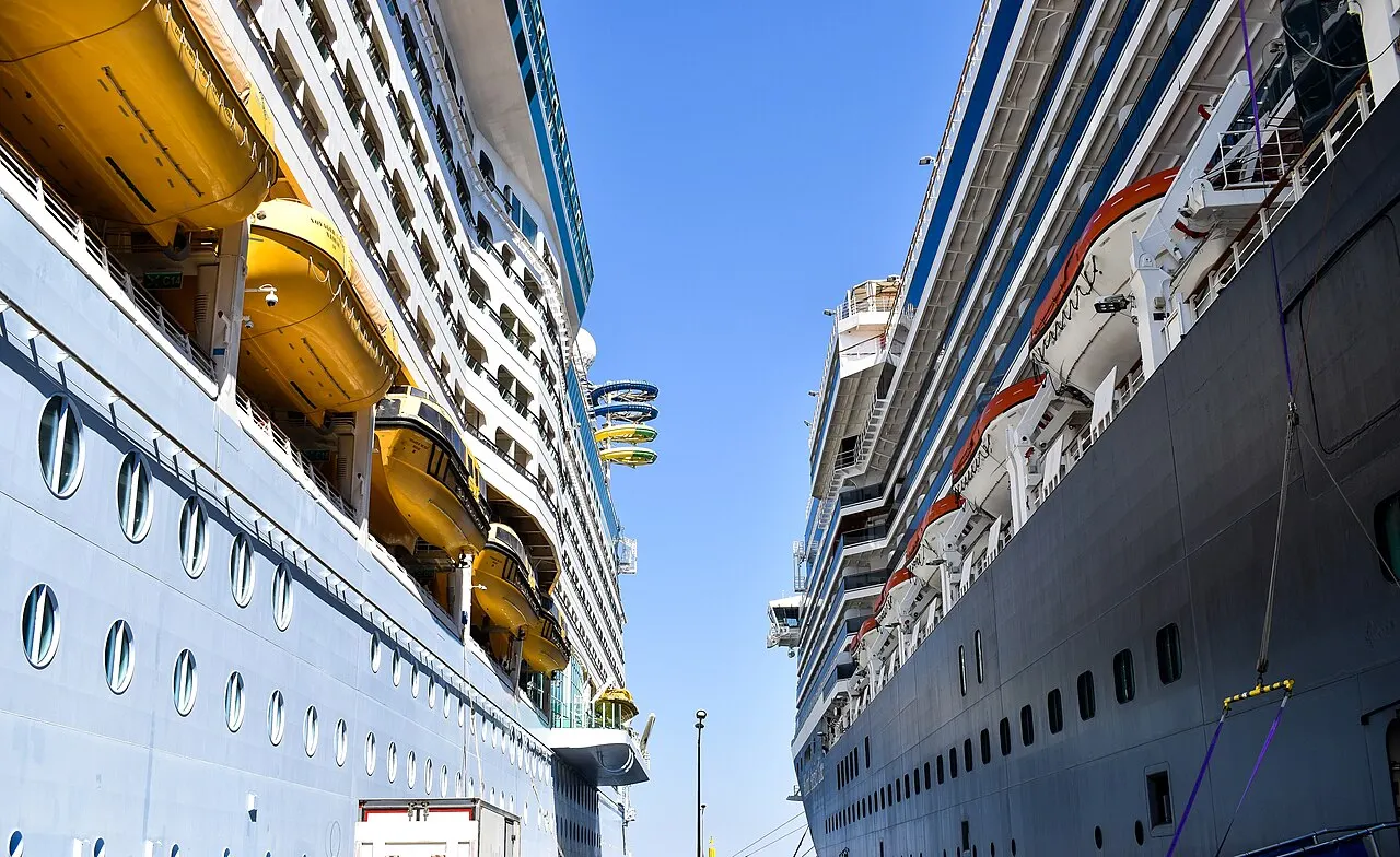 Foto: due navi da crociera affiancate alla banchina di Kuşadası, viste dallo spazio tra gli scafi, con scialuppe di salvataggio e file di finestre sui ponti.