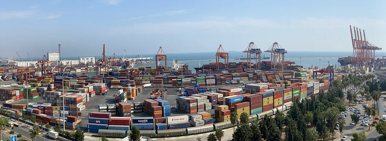 Foto: pile di container di molti colori nel piazzale del porto di Mersin. Sullo sfondo, gru di banchina e navi lungo l’acqua.
