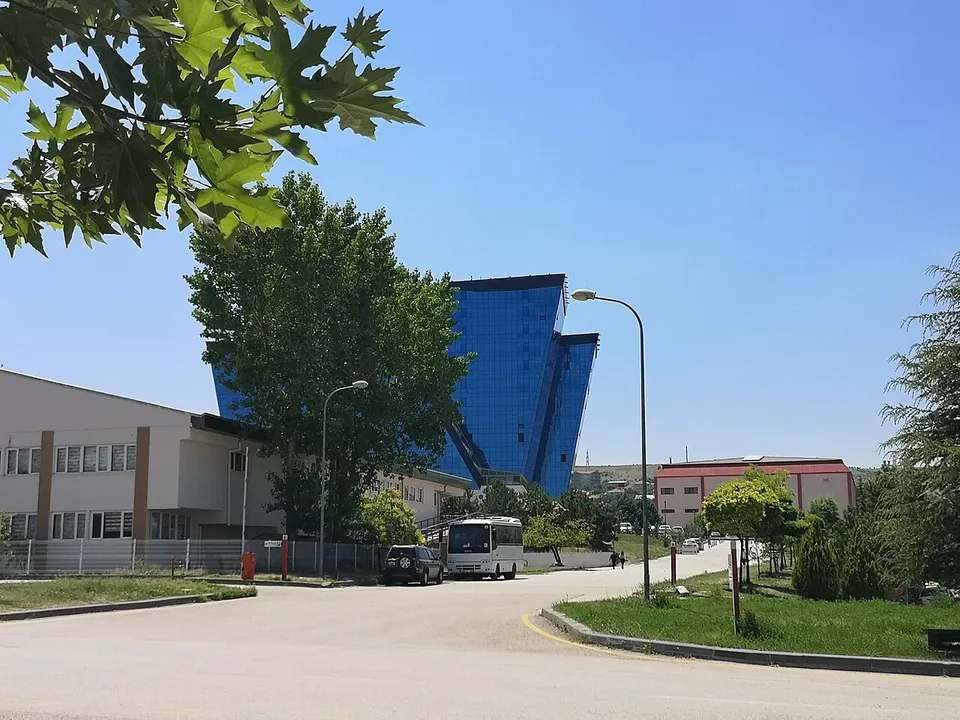 Ein Bürogebäude mit blauer Glasfassade hinter Bäumen und einer Campusstraße an einem sonnigen Tag, davor ein geparkter Kleinbus