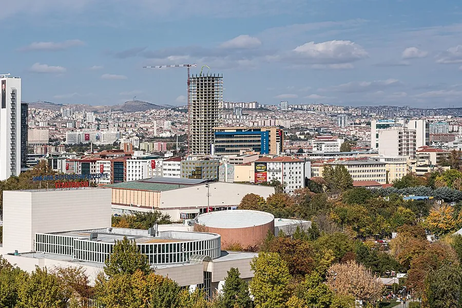 Ankara von einem erhöhten Standort aus gesehen: ein Park mit herbstlich gefärbten Bäumen vor einer langen weißen Halle, dahinter Wohnblöcke auf den Hügeln