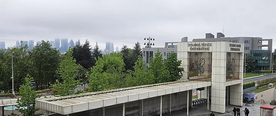 Ein Universitätscampustor mit flachem Dach und einem hohen Gitterpfeiler, Bäume und dahinter im Dunst die Hochhäuser von Maslak