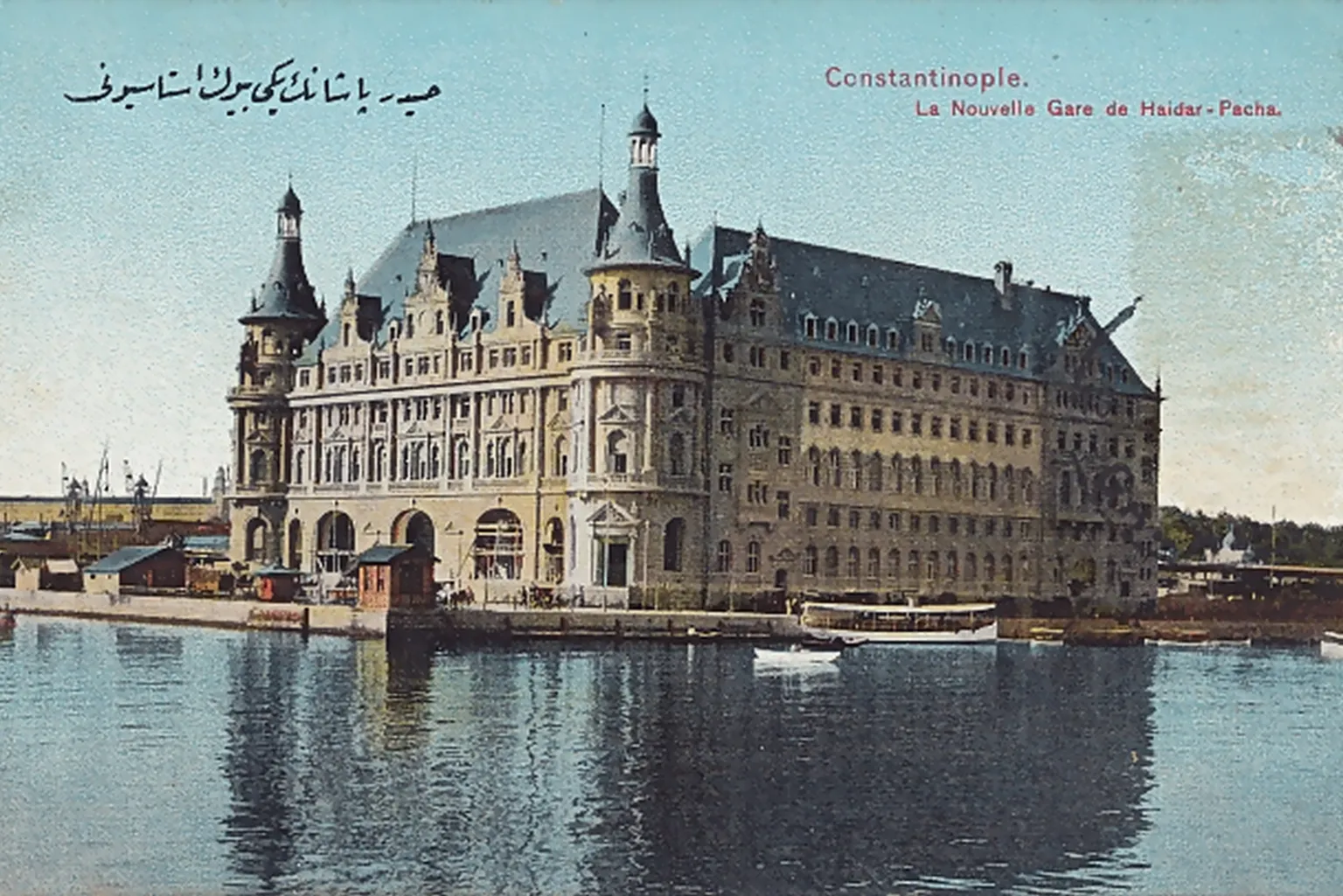 Colour postcard of Haydarpaşa station seen across the water: a long stone building with a steep slate roof, round corner towers with pointed caps and rows of arched windows, a small passenger boat at the quay, and the printed words «Constantinople. La Nouvelle Gare de Haidar-Pacha»