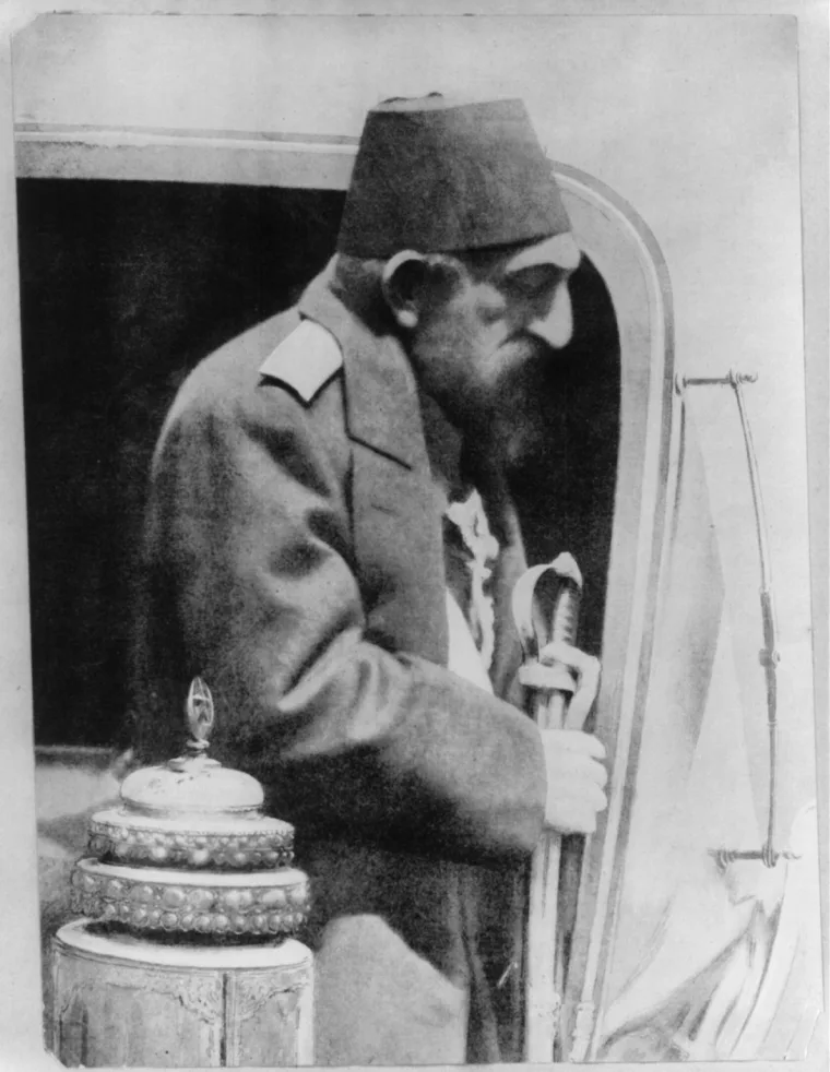 Black-and-white photograph of Abdülhamid II in profile: an older man with a beard, in a fez and a dark coat with epaulettes, stepping out of a carriage door and holding a sword, with an ornate lidded stand in the foreground