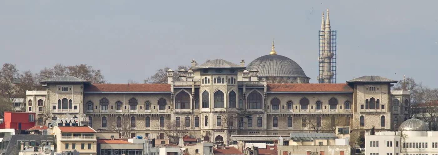 The same building today as İstanbul Erkek Lisesi: a long pale stone building with a red-tiled roof and tall arched windows in the middle, a grey dome and a minaret under scaffolding behind it, and houses in front