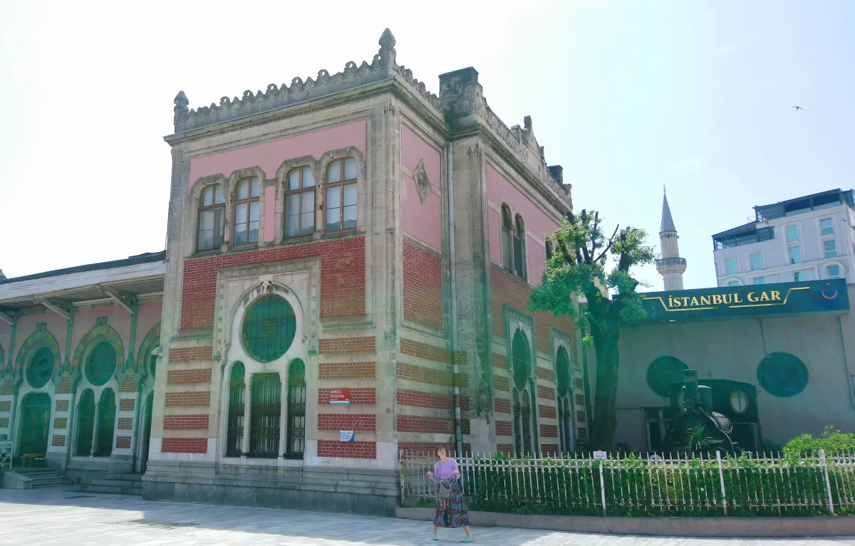 The side of Sirkeci station in Istanbul: a pale stone building with a pink upper wall, a lower wall of red and cream banded brick, arched windows with a round window in the middle, a small steam locomotive behind a fence at the right and a sign reading İSTANBUL GAR